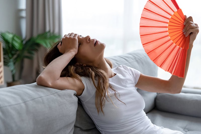 Why Is My AC Blowing Hot Air? Young adult woman is overheated, fanning herself on her couch.