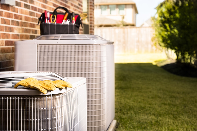 Why Do I Need AC Maintenance Now? Bag of repairman's work tools, gloves on top of air conditioner units outside a brick home. Service industry, working class.