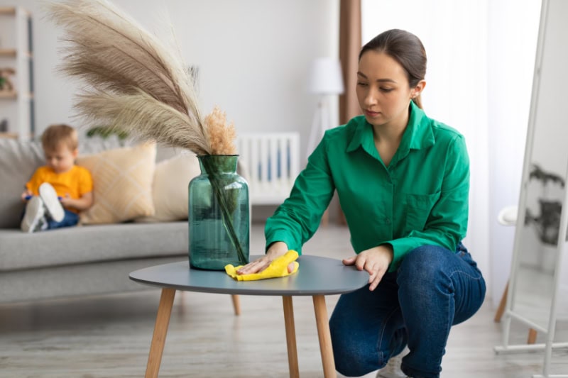 How to Reduce Dust in Your Home. Spring cleaning. Young mom dusting off coffee table and tidying up in living room while her toddler son watching cartoon on cellphone, sitting on sofa on background.