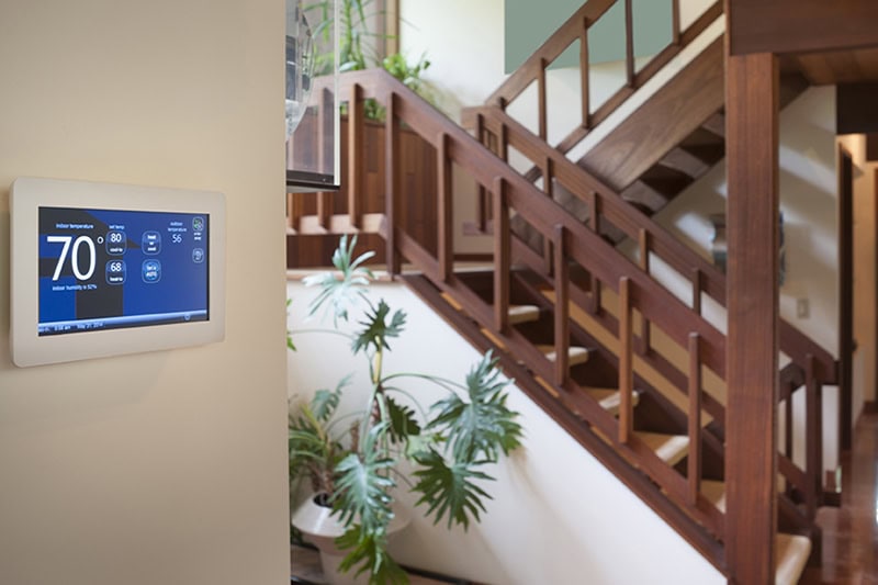 How a Smart Thermostat Can Save You Money During a Heat Wave. Photo of a thermostat mounted on a wall with a staircase in the background.