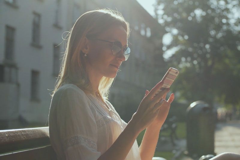 Video - Are Wi-Fi Thermostats Cost-Effective? Image is a photograph of a woman with blonde hair wearing sunglasses and looking at her phone while standing outside.